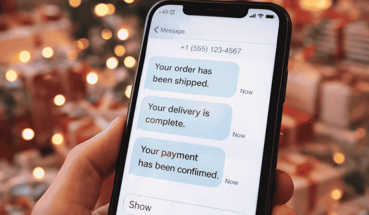 A hand holding a smartphone displaying three text SMS messages reading "Your order has been shipped," "Your delivery is complete," and "Your payment has been confirmed" against a blurred background of warm festive holiday lights.