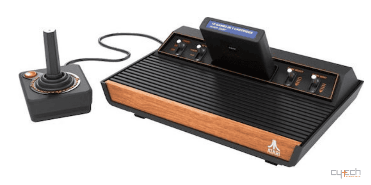 A classic Atari 2600 video game console with a woodgrain front panel, featuring an inserted multi-game cartridge and a connected wired joystick, set against a solid white background.