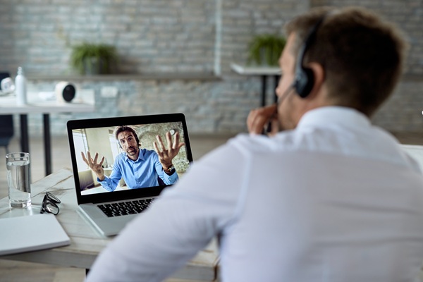 Custom WordPress Video Calling Platform by Cytech Over-the-shoulder view of an office worker wearing a headset, engaged in a video call on a laptop. The screen shows another man gesturing during a remote meeting, showcasing Cytech's custom-developed WordPress video conferencing service in use.
