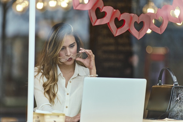 Modern Online Dating Platform – Scalable Cloud Infrastructure & User Experience A young woman looking attentively at her laptop while lowering her glasses in a cafe decorated with hanging paper hearts, representing an online dating and video communication platform